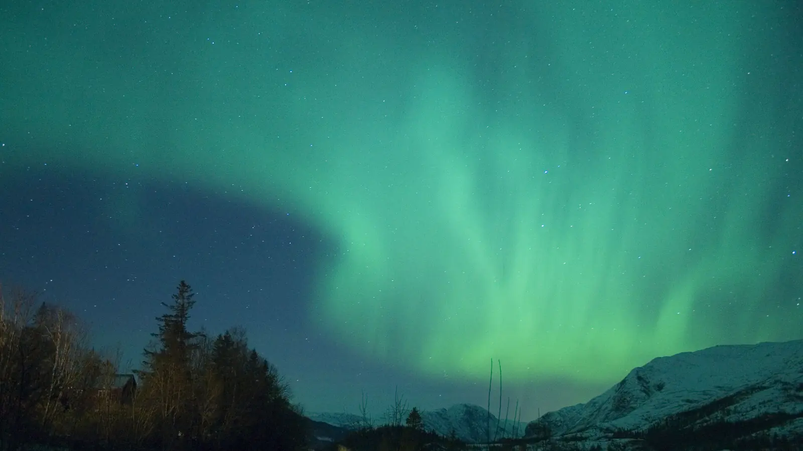 Green aurora curling over snowy mountains and a lit village at night