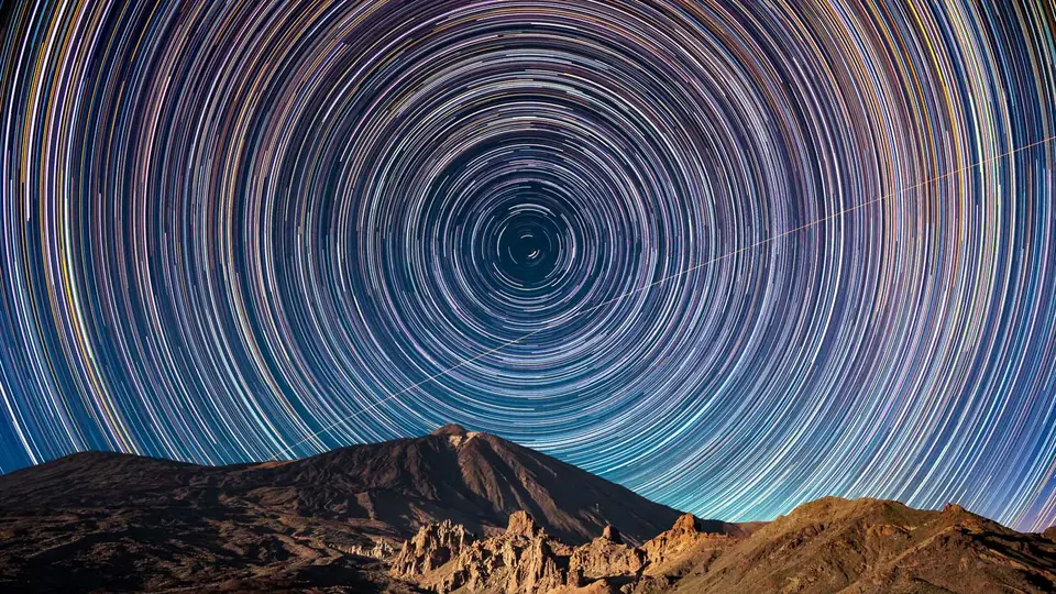 Star trails circling the celestial pole above a volcano, crossed by the straight line of a space station pass